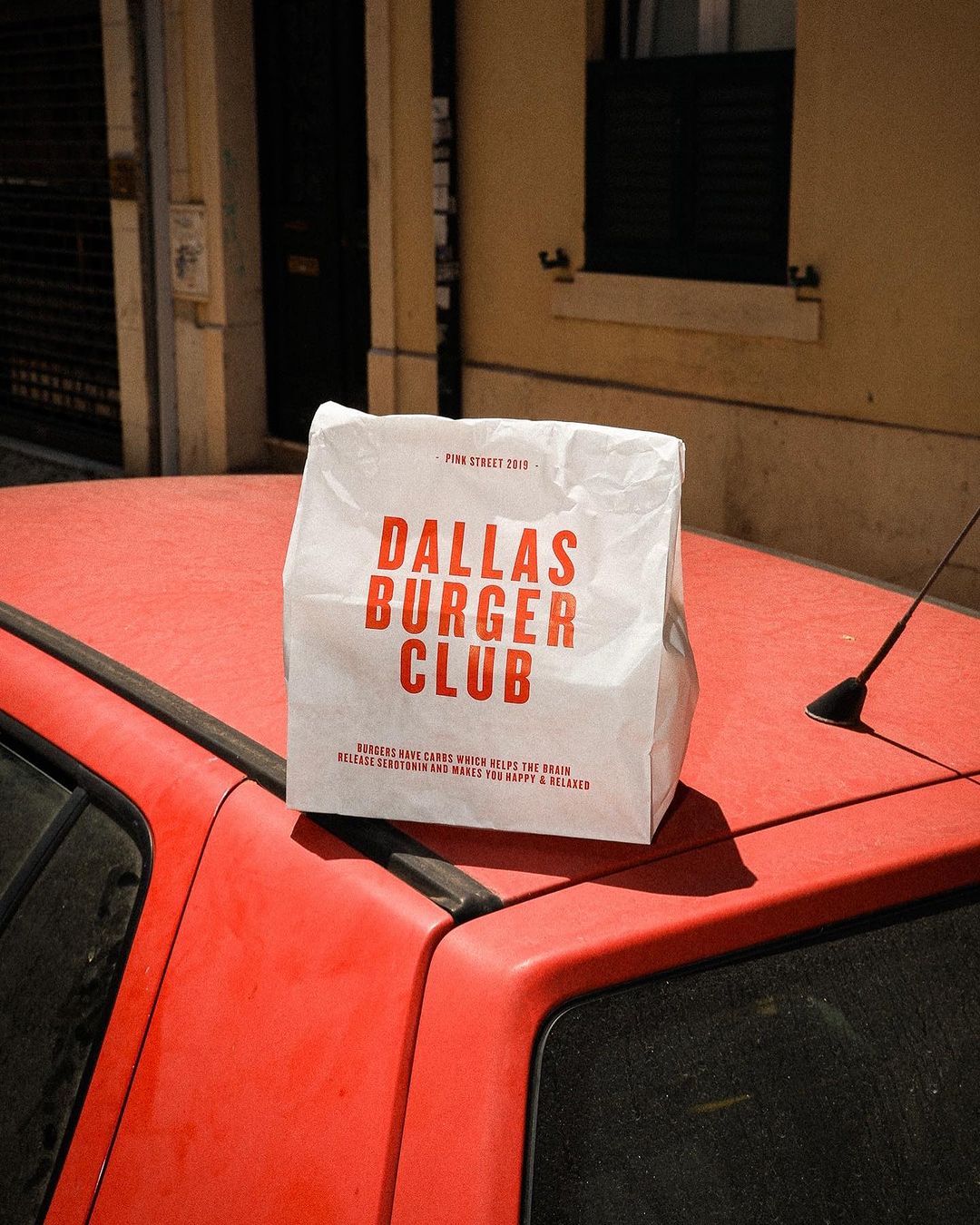 Branded takeaway bag from Dallas Burger Club placed on the hood of a red car in an urban setting.